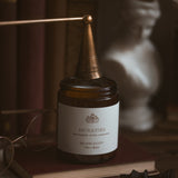 Jar candle labelled 'Antiquities' on a wooden surface, surrounded by books and a statue.