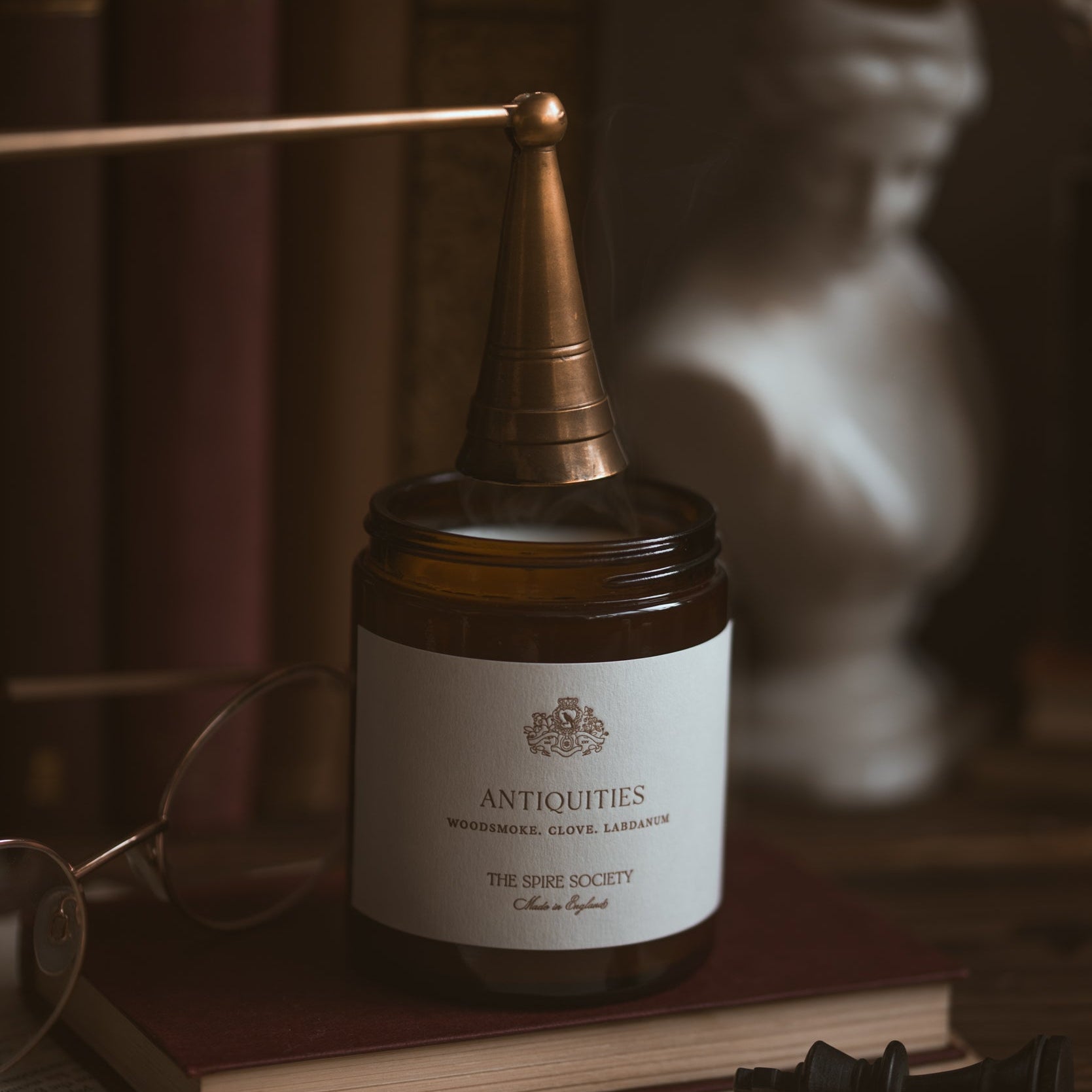 Jar candle labelled 'Antiquities' on a wooden surface, surrounded by books and a statue.