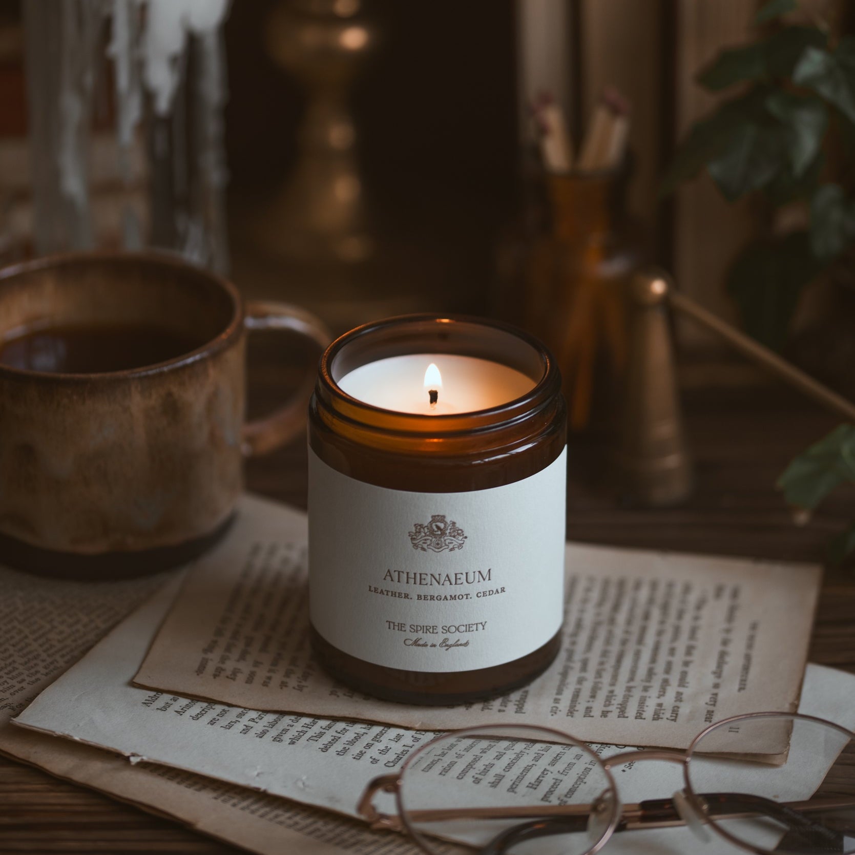 Candle in a glass jar with a label on a wooden surface with a cup and books in the background.
