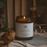 Candle in a jar labeled 'Ode & Elegy' on a surface with a blurred background