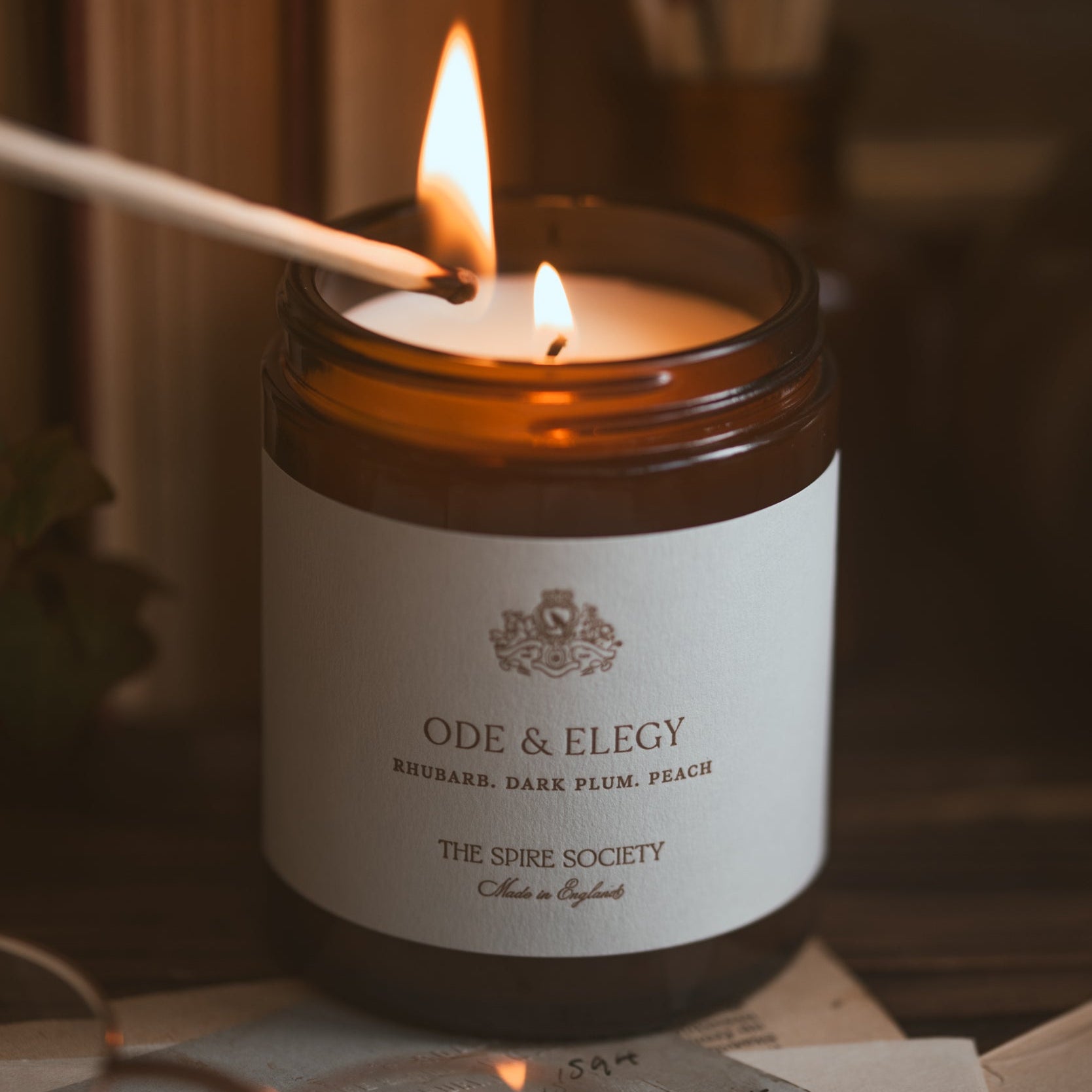 Ode & Elegy candle being lit with a match on a wooden surface.