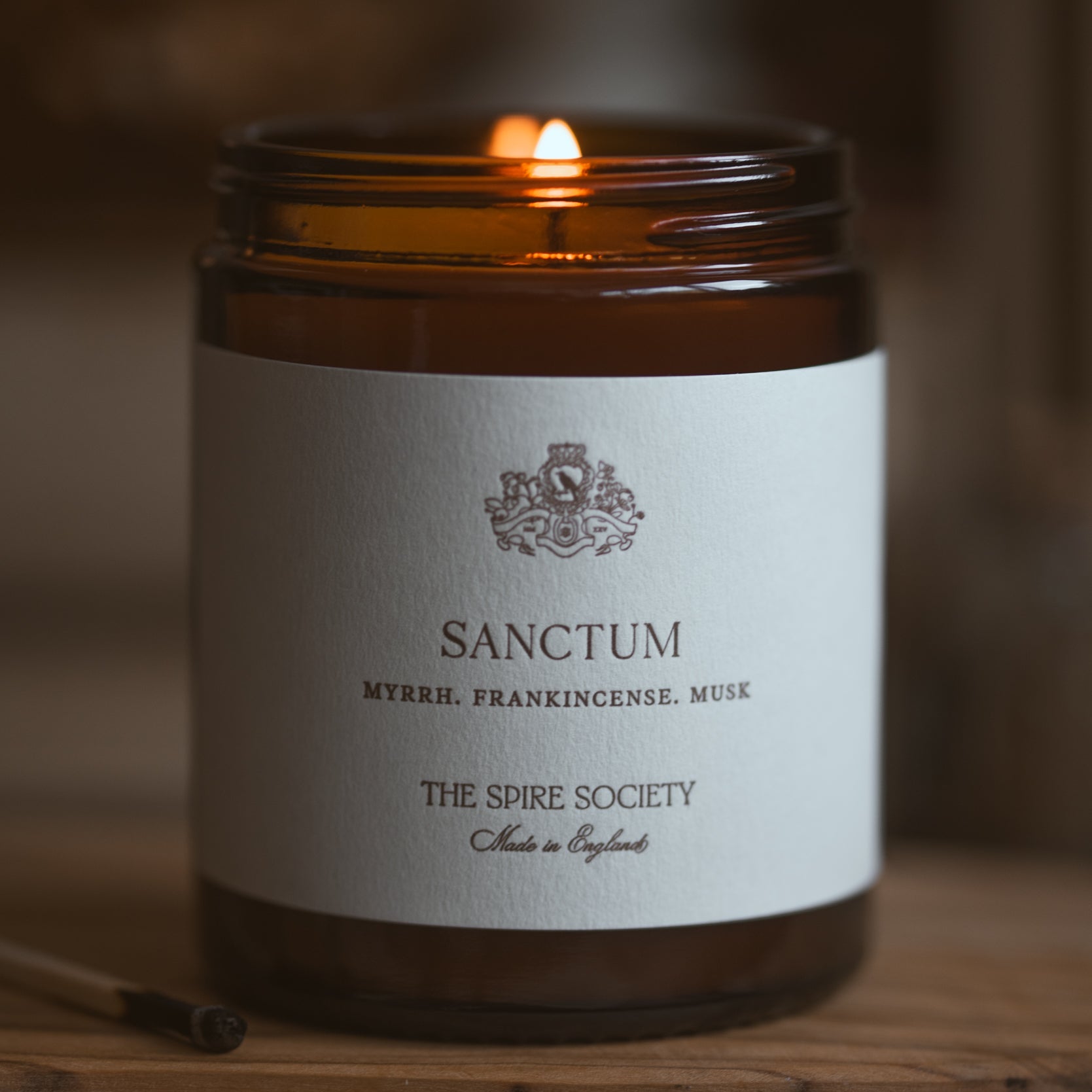 Candle in a brown jar labeled 'Sanctum' by The Spire Society on a wooden surface.