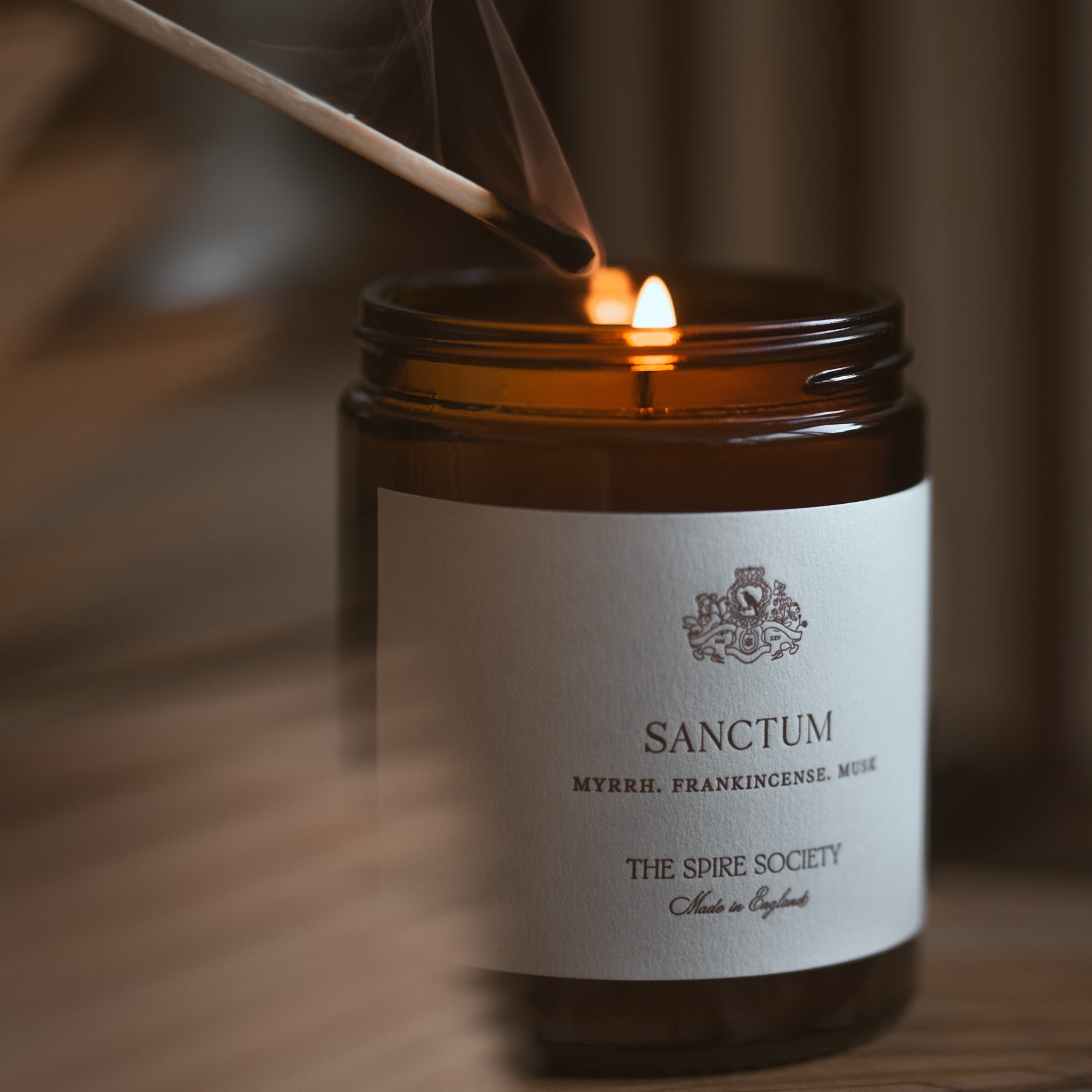 Candle being lit with a match, featuring the 'Sanctum' label on a wooden surface.