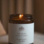Candle in a jar labeled 'The Quad' from 'The Spire Society' on a blurred background