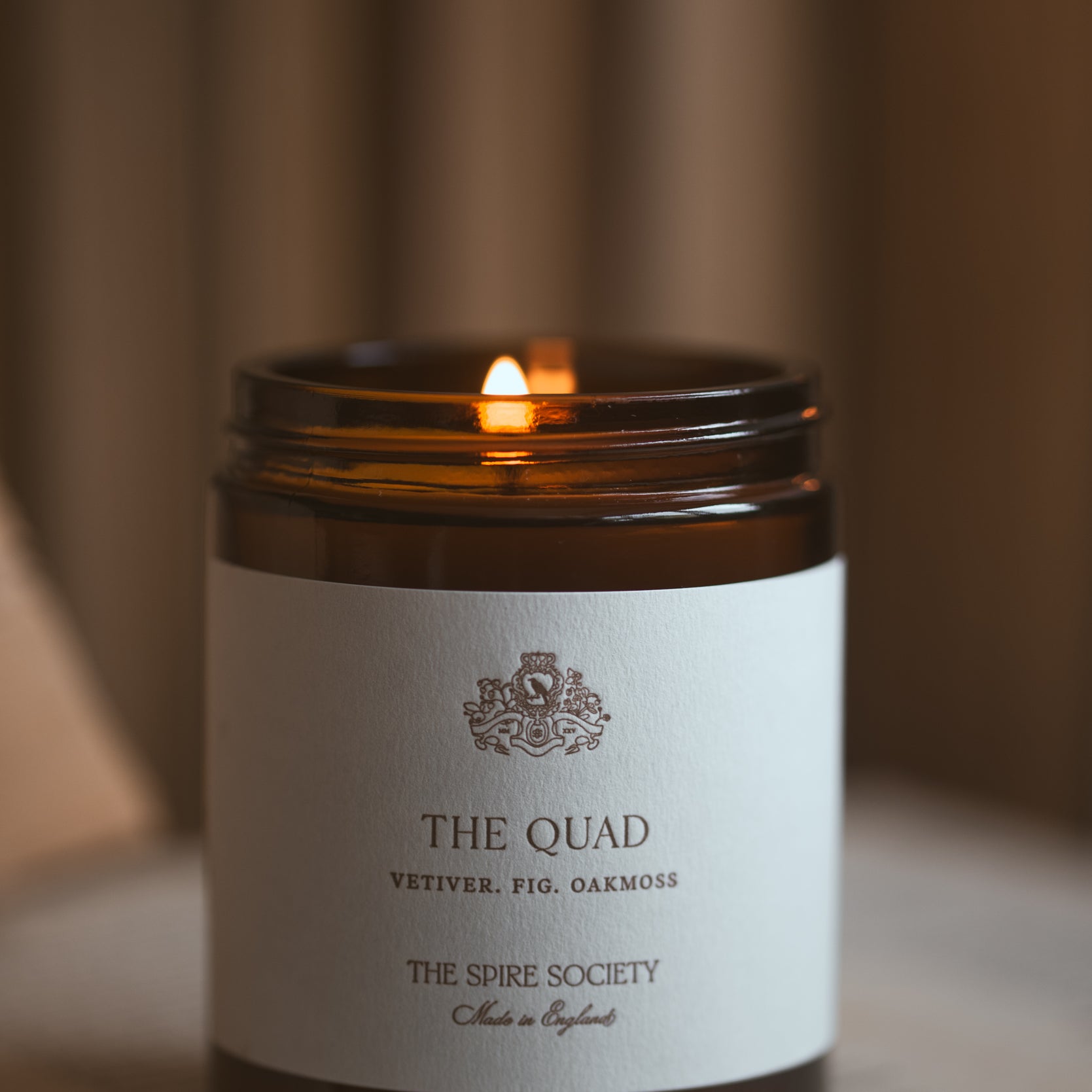 Candle in a jar labeled 'The Quad' from 'The Spire Society' on a blurred background